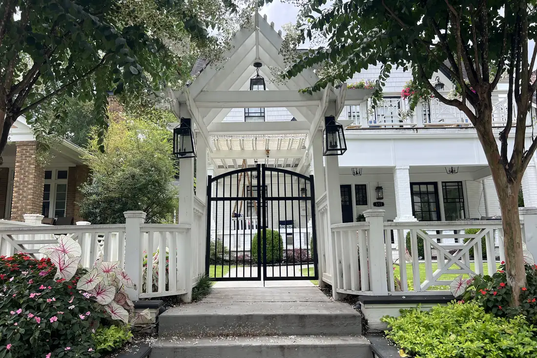 Custom black steel arched walk gate set under a white timber arbor with gas lanterns between two homes, built by FenceWorks of Georgia