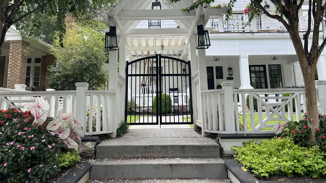 Custom arched ornamental steel entry gate under a white timber arbor with gas lanterns, designed and built by FenceWorks of Georgia